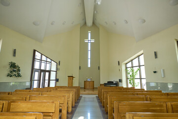 Empty hall for church services with aisle between two rows of wooden benches for parishioners and cross above pulpit of pastor - obrazy, fototapety, plakaty