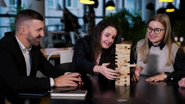 Jenga game played by adult group of people. Dark-haired woman pulls the little brick carefully to place it on top. Other players laugh and have good time.