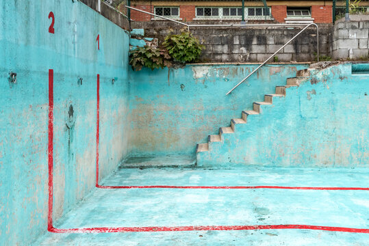 Abandoned public accessible swimming pool, focus on corner with stairs. The pool has a bright blue colour with red stripes.