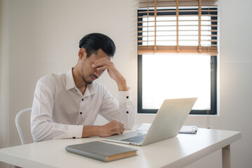 An Asian businessman in a white shirt sits at a desk with a computer holding his head in the stress of working in a white walled room.