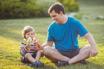 Cute and happy little child with his daddy in casual clothes eating ice cream together and sitting on lawn in the park