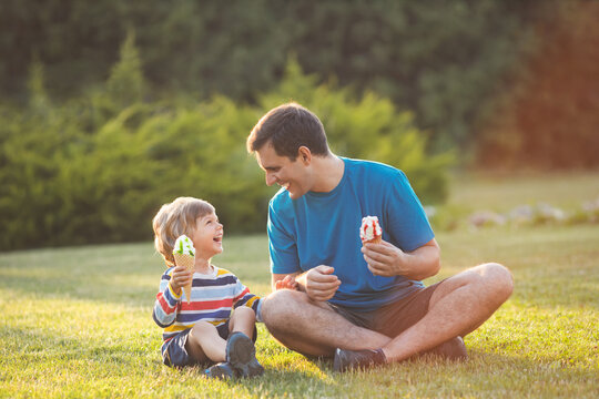 Laughing young father and little son haveing fun together and eating ice cream, sitting on grass in green park on summer day. Happy family concept