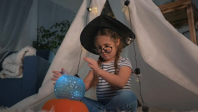 Cute happy child girl in top hat sits in tent, plays with magic lantern with glass ball.child fantasizes, dreams of witchcraft, fulfillment of desires.Happy child at home playing in room for christmas