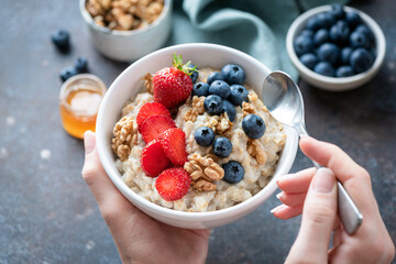 Oatmeal porridge bowl with berry fruits in female hands, closeup view. Healthy vegetarian breakfast...