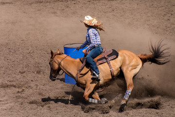 A Rodeo barrel racer is rounding a blue barrel. The cowgirl has a white hat, blue shirt and blue...