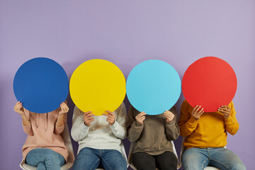 Young people hide their faces behind round colorful blue, yellow and red speech bubble banners that they are holding. Group of unrecognizable men and women trying to express their opinions anonymously