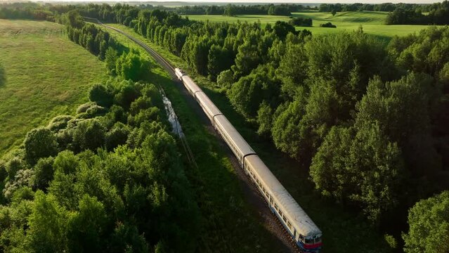 Passenger train on railway from forest, aerial view. Train with passenger cars rides along forests in spring. Drone view of an electric passenger locomotive with wagons in moving on railroad.