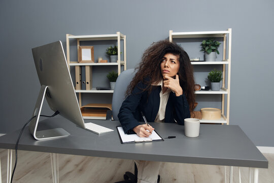 Thoughtful pensive tanned adorable curly Latin businesswoman in jacket ponder difficult project decision recline on hand looks up in office. Corporation leader lady uses modern computer in work