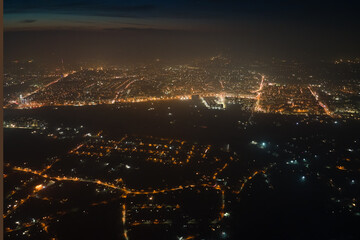 Aerial view from airplane window of buildings and bright illuminated streets in city residential area at night. Dark urban landscape at high altitude