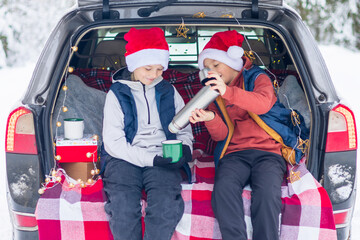 Naklejka premium two children friends boys in Santa hats traveling and having fun in trunk car in snow winter forest, drinking tea from thermos, talking and getting ready to celebrate Christmas or New year together