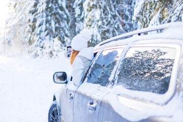 teenage girl in white sweater, vest and white knitted hat in car window in snowy forest take selfie photo on mobile phone, concept winter local travel during Christmas or New Year holidays vacation