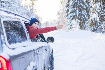 teenage cute boy in white sweater, vest and white knitted hat in car window in snowy forest having fun, concept of winter local travel during Christmas or New Year holidays and vacations