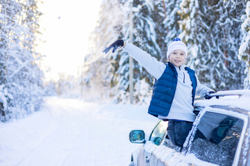 teenage cute boy in white sweater, vest and white knitted hat in car window in snowy forest having fun, concept of winter local travel during Christmas or New Year holidays and vacations