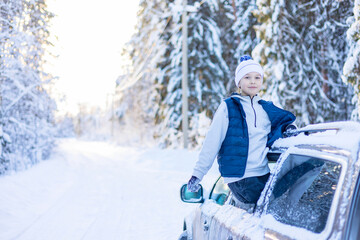 teenage cute boy in white sweater, vest and white knitted hat in car window in snowy forest having fun, concept of winter local travel during Christmas or New Year holidays and vacations