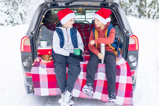 Two Children Friends Boys In Santa Hats Traveling And Having Fun In Trunk Car In Snow Winter Forest, Drinking Tea From Thermos, Talking And Getting Ready To Celebrate Christmas Or New Year Together