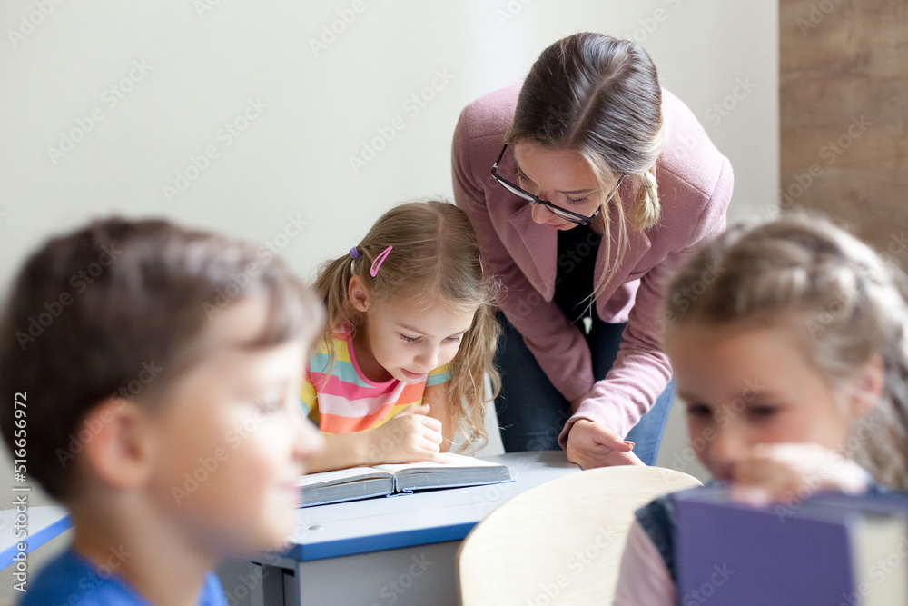 Wall mural School teacher helping pupils studying, reading book. Lesson for kids in elementary college. Children sitting at desks in classroom. Woman guiding in-person, teaching primary students. Back to school