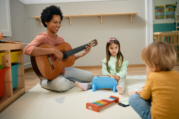 Happy black kindergarten teacher playing acoustic guitar to group of kids.
