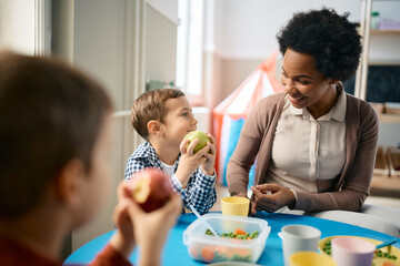 Happy black teacher and small boy talk during lunch time at kindergarten.