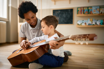 Little boy learns to play acoustic guitar with help of his preschool teacher.
