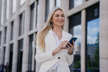 Purposeful young caucasian blonde woman in white suit standing outdoor with phone on summer shiny day. Happy student swedish girl going to call her boyfriend. Cute smiling Italian pretty woman.