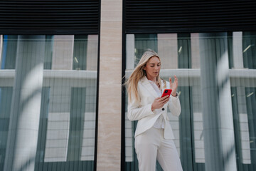 Pretty young lawyer in white suite standing outdoors against contemporary building with large windows making video call. Italian confident entrepreneur arguing with partner, agent convinces client.