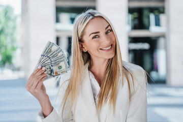 Excited blonde caucasian woman in white suite holding US dollar banknotes smiling wide, satisfied...