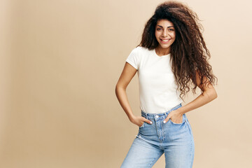 Overjoyed woman with afro hair in white t-shirt, smiling at camera, hold hands on pocket, say Yeah, isolated beige background. Copy space clothing fashion brands, free place for your ad