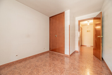 Empty room with built-in wardrobe with wooden doors and matching carpentry, off-white painted walls and reddish stoneware floors