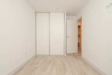 Empty bedroom with light wood flooring, plain white painted walls and built-in wardrobe with white sliding doors