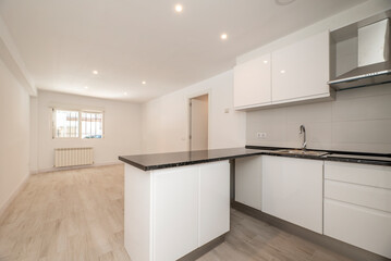 Open plan kitchen of an empty apartment with plain white painted walls with handleless cabinets and black quartz countertop