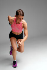Determined muscular female athlete workout, raising leg and doing stretching exercises. Sport woman in sportswear training indoors, doing fitness aerobics, gray background.
