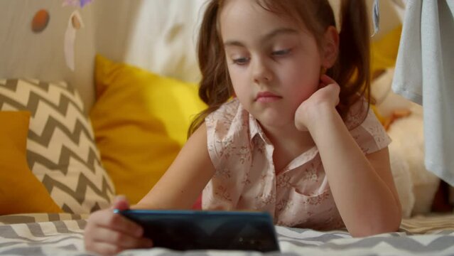 Little cute girl uses a smartphone while lying on the floor in a tent at home. child watches the Internet or films, spends holidays at home.