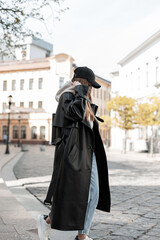 Fototapeta na wymiar Fashionable beautiful street hipster woman in stylish urban casual clothes with a fashion black long leather coat and cap walking in the city and wearing a hood