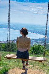 A young girl sits on a swing in the mountains overlooking the sea coast and the resort town