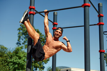 Athletic young man hanging from the bars at the calisthenics gym outdoors smiling - obrazy, fototapety, plakaty