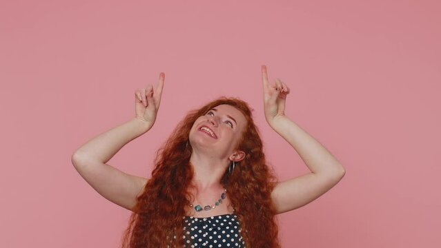 Lovely redhead woman showing thumbs up and pointing up empty place, advertising area for commercial text, copy-space for goods promotion. Young ginger girl. Indoors studio shot on pink wall background
