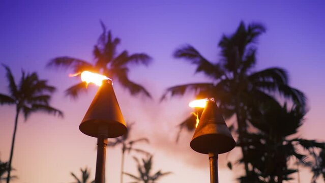 Tiki torches held by tiki gods lining the hut of an outdoor Polynesian luau at night. Dramatic pink velvet purple sunset sky on motion background of the bright fire flames blowing from island torches