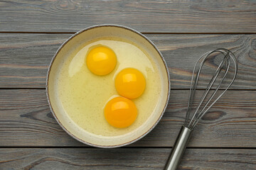 Whisk near bowl with eggs on wooden table, flat lay