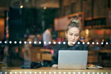 through the window glass shot of mid adult asian business woman working in coffee shop using laptop computer