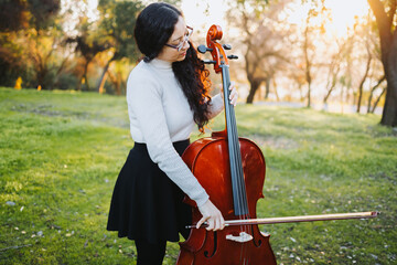 Young standing brunette woman with glasses playing cello at sunset in the park, on a green grass. © padnob