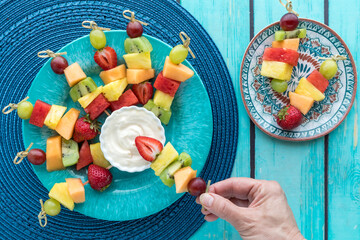 A tray of fresh fruit skewers with a hand dipping one into a yogurt dip.