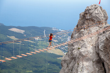 A young girl walks across a suspension bridge on an abyss in the mountains against the backdrop of the sea on a sunny summer day.Safe adventures while traveling, healthy image zhini concept