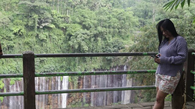 Asian woman replies to chats with her smartphone against the background of the tumpak sewu waterfall in Indonesia