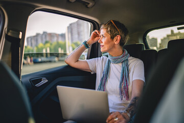 one mature woman caucasian female sitting on the back seat of the car thinking while looking to the side and working on laptop computer in summer day with short gray hair modern manager copy space