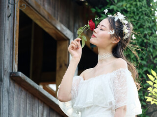 Beautiful fairy lady in white wedding dress and garlands of flowers, eyes closed with red rose in...