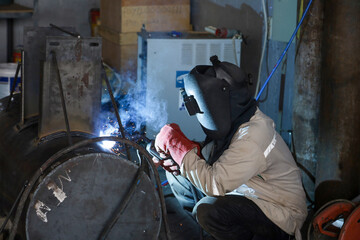 A young man welder in brown uniform, welding mask and welders leathers, weld metal with a arc...