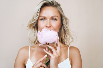 Beauty portrait of blonde hair young woman with pink peony in hand isolated on beige background