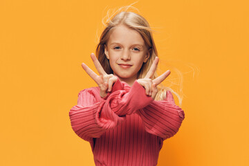 a cute, happy school-age girl stands on a yellow background in a pink sweater and shows a victory sign with her fingers smiling broadly