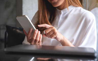 Closeup image of a young woman holding and touching on mobile phone screen with laptop on the table
