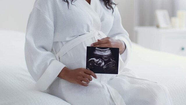Baby first photo. Unrecognizable black pregnant lady in bathrobe holding sonogram and caressing her belly, slow motion